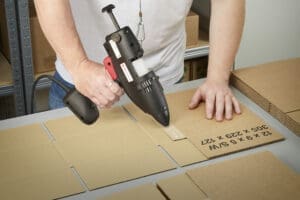 Man using an heavy industrial 43mm glue gun to apply hot melt to a cardboard box tab.